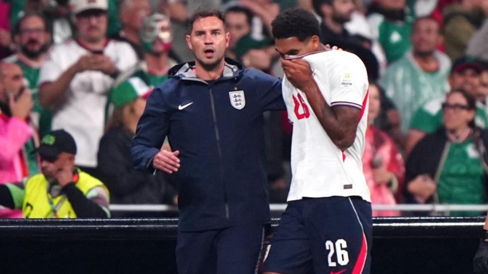 England defender Jarell Quansah walking off the pitch after he was shown a straight red card in the Three Lions win over Mexico in Mexico City. He has his head in his hands and and is being consolidated by a member of the coaching staff.