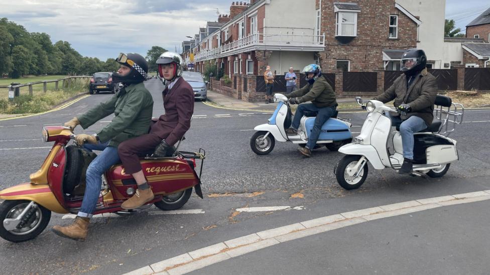 Three moped scooters going round a corner. The first scooter is red and gold and being ridden by a man wearing a green jacket and a black helmet with a teenage boy wearing a dark purple suit and a silver helmet sitting on the back. Behind are two scooters - one white and one blue and white - being ridden by men wearing dark green jackets and blue jeans and helmets.