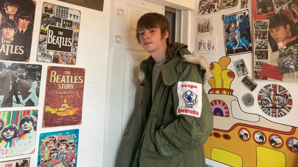Teenage boy with brown hair going over his ears is wearing a green parka jacket with a cream edge on the hood. Travis is stood in front of a white bedroom door with The Beatles posters on the wall to the left and right of him. Behind Travis is a colour photo of a yellow submarine featured on one of The Beatles albums.