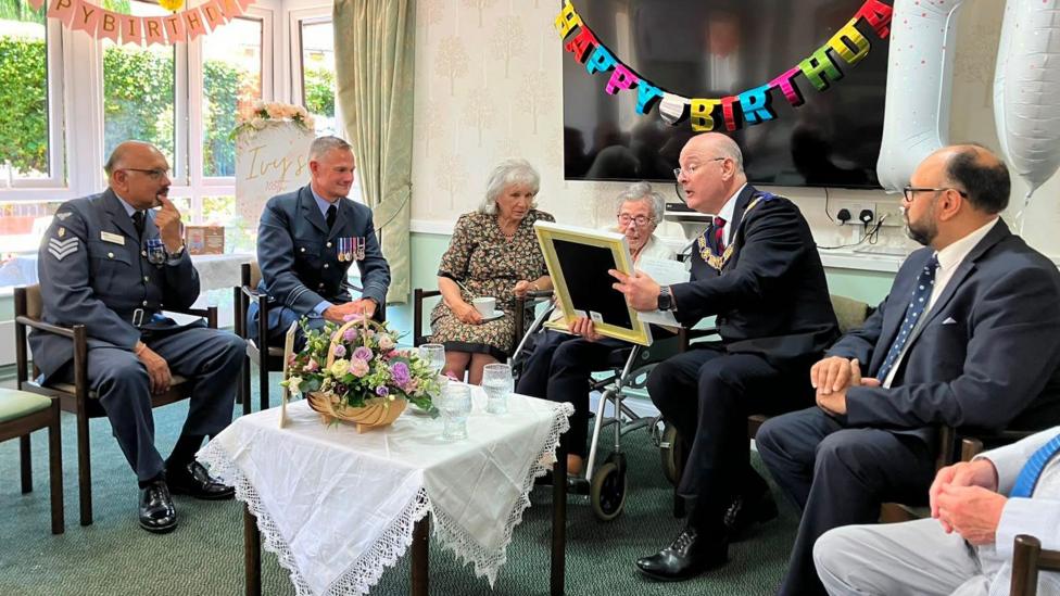 An elderly woman is sat in the middle of six people as she receives an honour from a man sitting next to her.