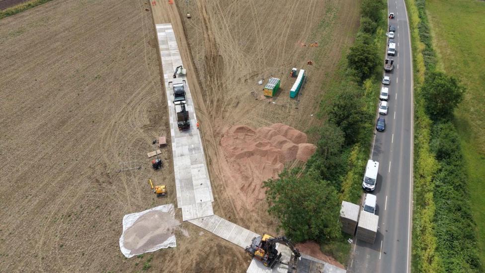 An aerial shot of a field with a metal track next to a road, with vehicles parked along the road. There is sand in the field. There are trees and bushes along the road.