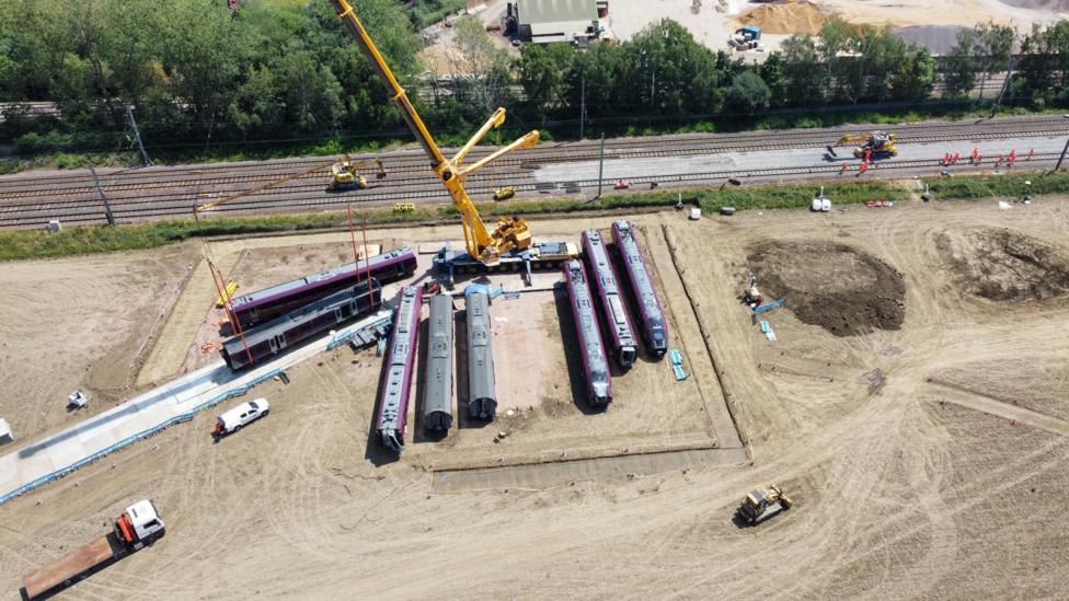 A crane has lifted train carriages off a track on to a field which has a temporary road running into it.
