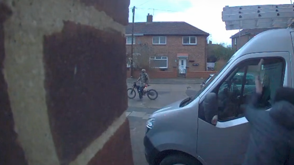 An image from a doorbell camera shows a person is sitting on a dirt bike on a road in the background while in the foreground somebody else hitting the passenger side window of a parked silver van. The attacker is using a weapon, believed to be an axe. It is happening in a housing estate with two-storey red-brick houses visible on the other side of the road.