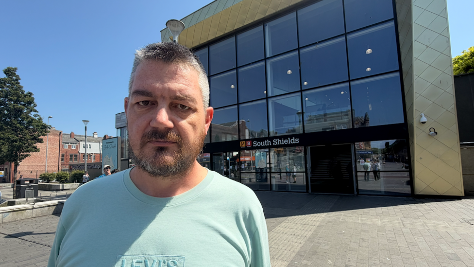 Mark Berriman is standing outside South Shields Metro station on a sunny, cloudless day. He has short cropped grey hair and a brown and grey beard. He is wearing a greeny-blue t-shirt with a Levi's logo embossed on the front in a slightly different shade. The station is a tall building with the sides and top clad in a gold colour containing symmetrical diamond shapes and surrounding huge darkened windows in a continuous pane of squares marked out with a thin black partition lines. These are arranged in three rows of six above a glass full-width entrance which includes automatic sliding doors.