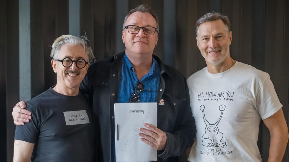 Davies stands in the middle of Cumming and Morrissey, with his hand around Cummings' shoulders. Davies holds the series script in one hand. Morrissey wears a white t-shirt with a graphic on, and has short grey hair. Cumming wears black round glasses and wears a black t-shirt.