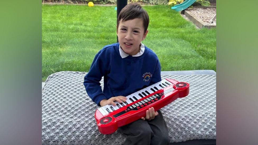 A boy wearing a blue jumper and grey trousers. He is holding a children's toy keyboard and there is grass behind him