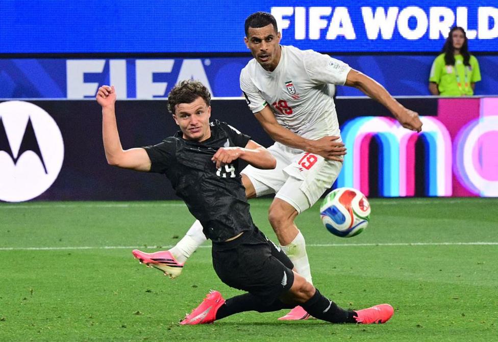 A footballer in a black New Zealand strip flies into a tackle on the Iranian number 19 in a white kit