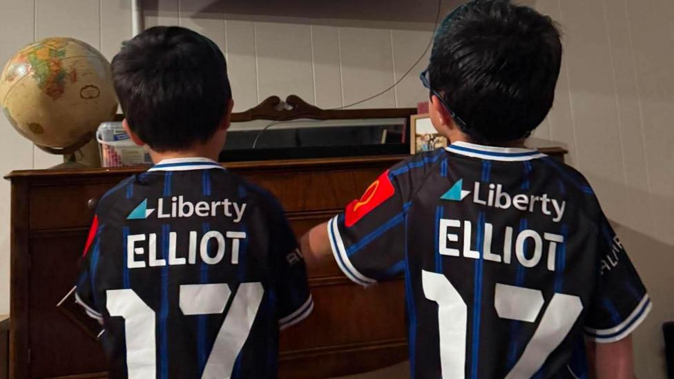 Two little boys stand side by side with football strips with Elliot 17 on the back of them