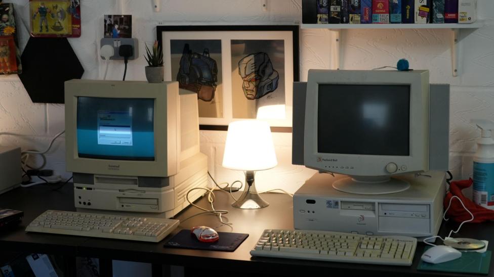 Two retro computers side by side on a desk each with a mouse and keyboard. In between them is a lamp which is turned on.