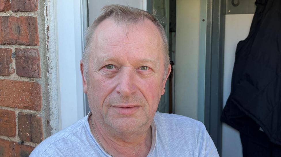 Picture of Mr Sampson stood in his doorway. He wears a grey crew neck t-shirt and a silver chain. He has blue eyes and short silver hair. He has a short stubbled beard.