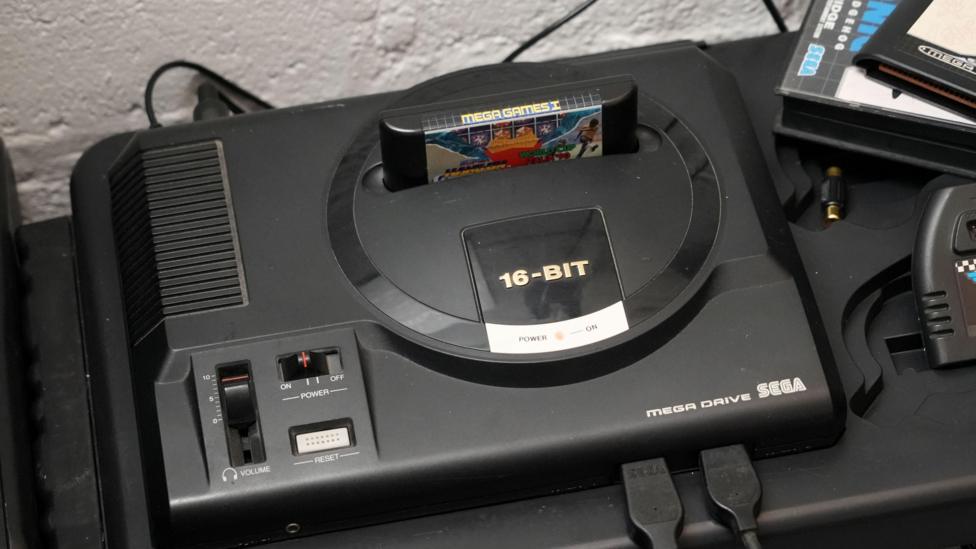 A black Sega Mega Drive is shown on a black table, with a game cartridge inserted into the machine.