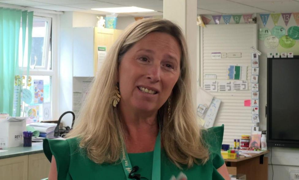 Headteacher Kate Davies is wearing a green sleeveless dress with a lighter green Pines Primary School lanyard round her neck. She is standing in front of a large window with the light streaming through it despite the vertically slatted blind that's pulled partially across it.