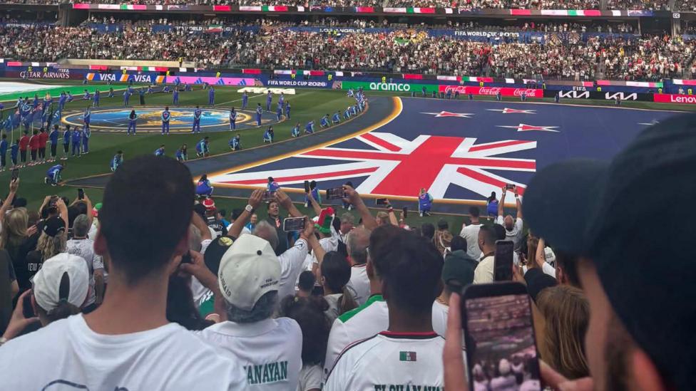 A view of a packed football stadium through a group of fans towards a massive New Zealand flag on the pitch