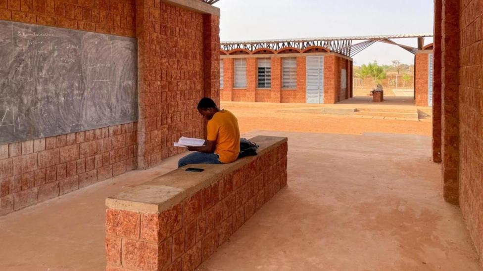 A young man sits alone reading a book sitting on a low wall outside a school in Tanzania. The school's walls are made of large red blocks and the corridors between the different school, building are left option to help with the air flow.