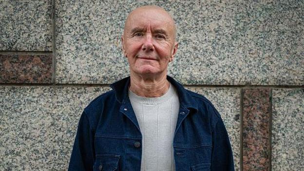 Irvine Welsh, a bald man in a blue denim jacket and light grey jumper, smiles in front of a marble wall.