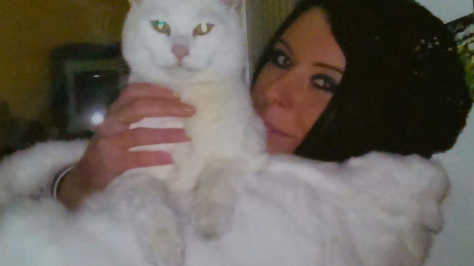 A woman with dark hair wearing a fluffy white jumper, holds a white cat on her chest.