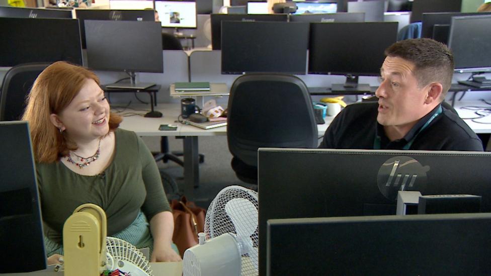 Two people in an office sit facing each other in an open-plan office, talking across computer screens. 