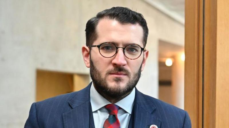 Paul Sweeney - a man with short dark hair, glasses and a beard, wearing a dark blue suit.