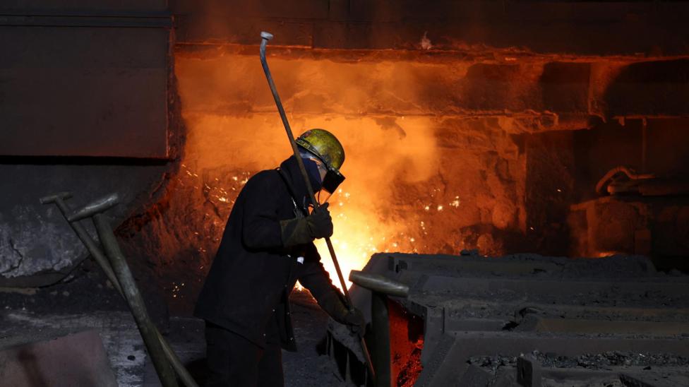 A steel worker standing in front of a furnace with his outline dramatically lit up by the flames behind him. The man is wearing protective overalls, a helmet and goggles, and holding what looks to be a long metal rod or pipe.