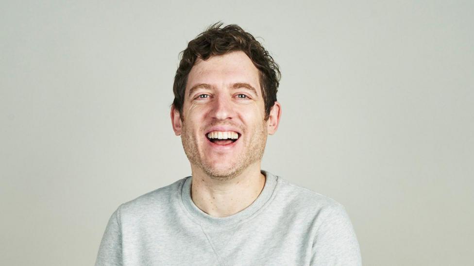 Elis James with short brown hair and stubble, wearing a grey jumper, smiles at the camera