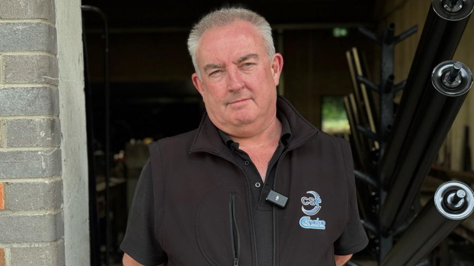Michael McGrath, a man with short, spiky grey hair, stands at the entrance of a workplace, looking directly at the camera. He is wearing a black polo shirt under a black body warmer.