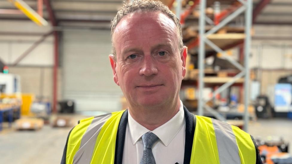 Stephen Kelly, a man with short, curly fair hair, looks at the camera in a factory or warehouse setting. He is wearing a yellow high-viz vest over a dark suit, white shirt and blue tie.