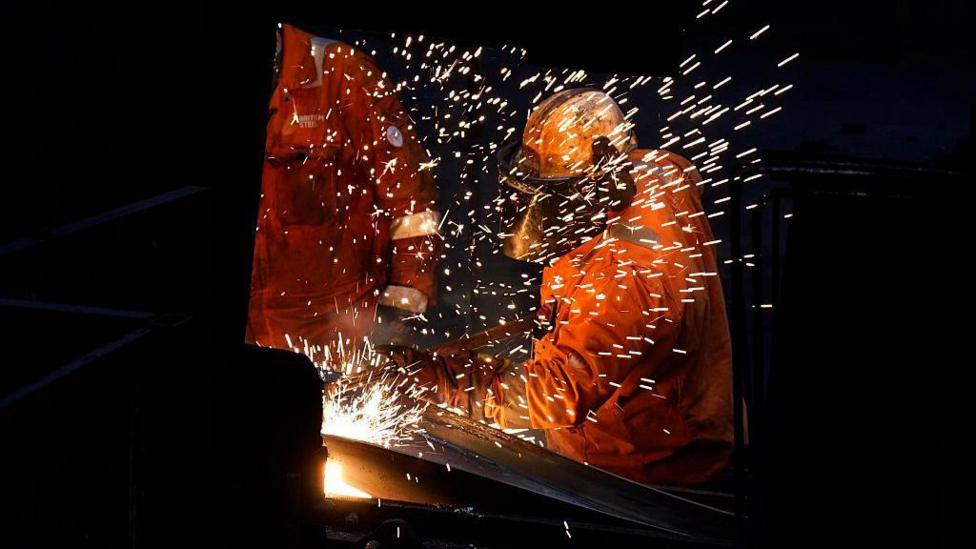 Sparks fly as workers wearing hi-vis orange jackets and helmets work with metal at a steel factory.