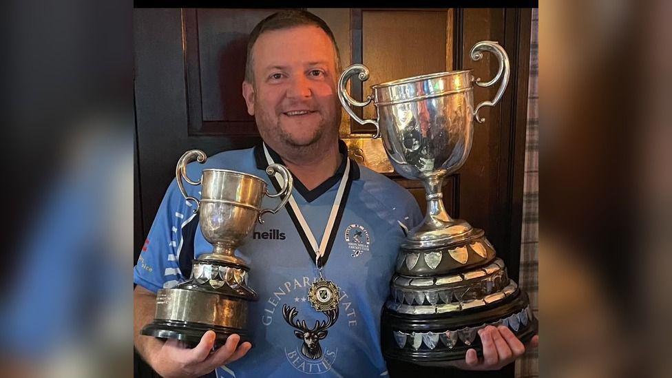 A cricketer, in a blue uniform, holds two trophies, one in each arm. A medal is around his neck and he is smiling.