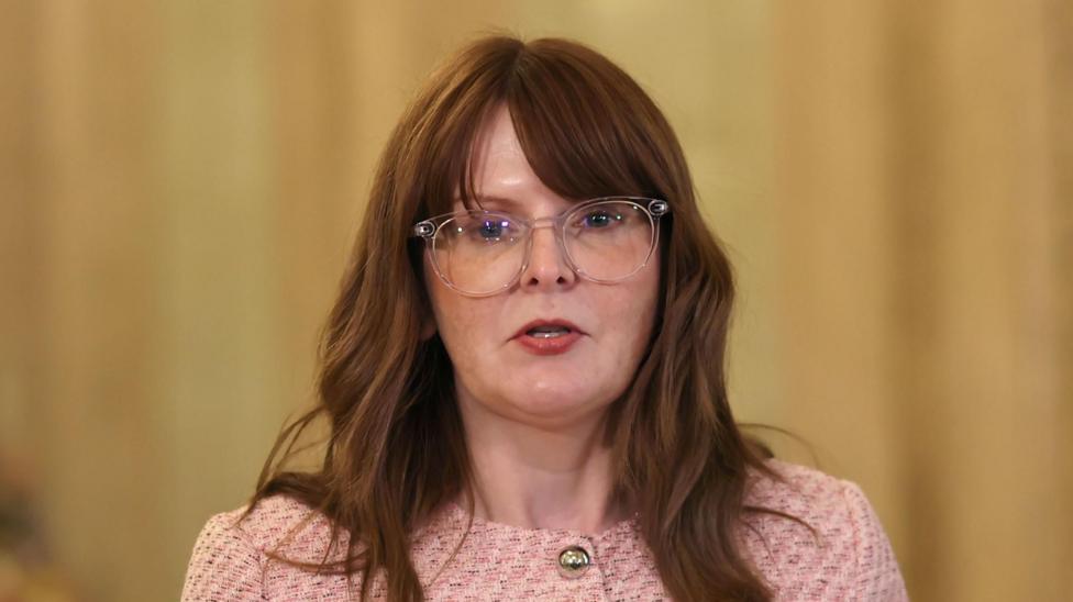 Caoimhe Archibald, a woman with reddish brown hair, wearing glasses and a tweed pink jacket.