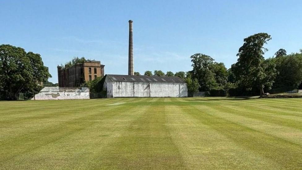 A green grassed grass cricket field stretches towards an old industrial building with a high chimney stack. The sky is blue. A number of trees are on either side of the building.
