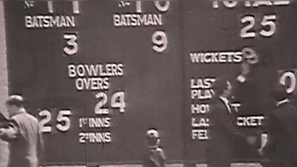 A screenshot from a video of a 1969 BBC film of Ireland playing against west Indies in cricket in which a number of people stand around an old style cricket scoreboard. The image is black and white
