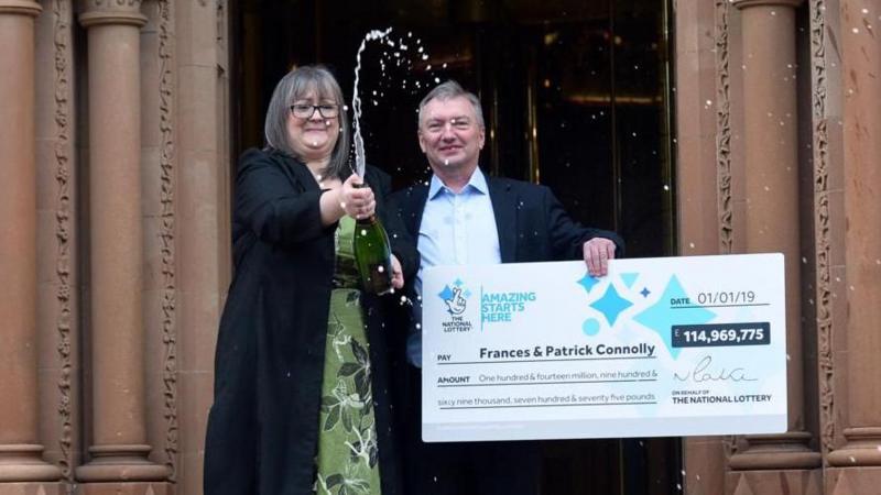A woman pops open a bottle of champagne as she stands on stpes alongside a mn holding an oversized cheque. He is wearing a blue shirt and jacket. She is wearing a long black coat and green dress. They have won the lottery.