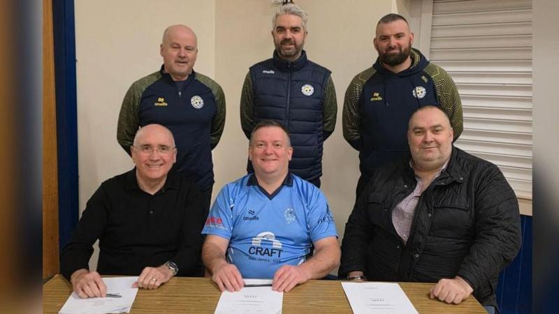 Three men sit at a table with paperwork in front of them. Behind them are three men who are standing . The standing men are wearing the tracksuits of a football club. The man in the middle sitting is wearing a cricket club shirt.