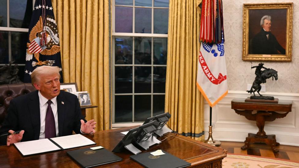 President Donald Trump, a man with short, blond hair, sitting at a wooden desk in the Oval Office speaking to the media. On the wall to his left there is a gold-framed painting of President Andrew Jackson. Trump is wearing a dark suit jacket, a white shirt and a purple tie. There are document folders on his desk. Behind him there are flags and large windows draped with gold-coloured curtains.