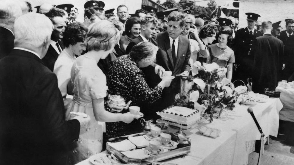 A black and white photo of US President John Fitzgerald Kennedy sharing a lunch with his Irish family during his visit in Dunganstown, County Wexford in 1963. He is wearing a dark suit and tie and is standing at a long table, surrounded by women, while uniformed police stand guard in the background. A woman is handing him a cup of tea. There is a tea tray, a large white cake and flowers on the table.