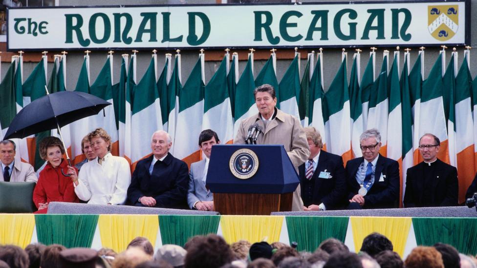 The late US President Ronald Reagan addressing a crowd in Ballyporeen, County Tipperary in June 1984. He has short black hair and is wearing a beige raincoat. A large pub sign saying The Ronald Reagan is behind him, along with a line of Irish flags. Several people are on a stage behind him including his wife Nancy who is being sheltered by a woman holding an umbrella.
