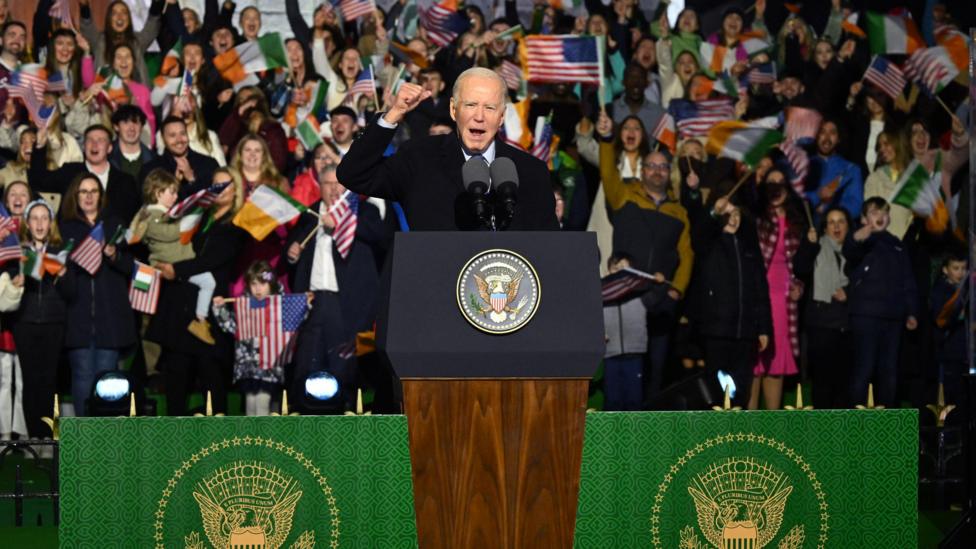 Joe Biden stands at a lecture addressing a large crown of well-wishers in Ballina, County Mayo, in 2023. He had short, white hair and is wearing a long black coat over a shirt and tie. People in the crowd behind him are waving US and Irish flags.