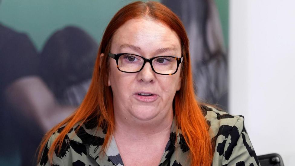 A woman with red hair and a khaki and black shirt, wearing dark-rimmed glasses.