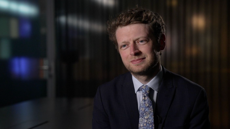 Connor Rand smiles, wearing a black suit, white shirt and floral tie, as he is interviewed in an office room
