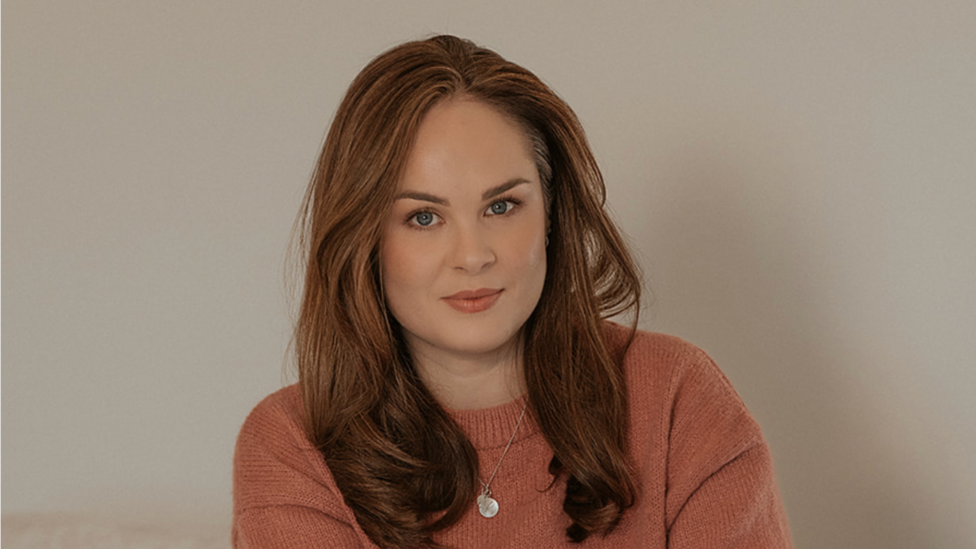 Headshot of a woman in her 30s, looking at the camera with a blank background. She has long ginger hair and is wearing a burnt-orange jumper and a silver necklace.