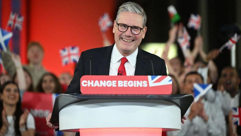 Sir Keir Starmer smiles from behind a red podium reading