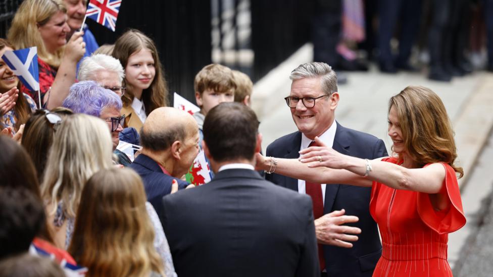 Prime Minister Keir Starmer and his wife Victoria Starmer in red dress with arms outstretched greet well-wishers as they arrive at Downing Street in London