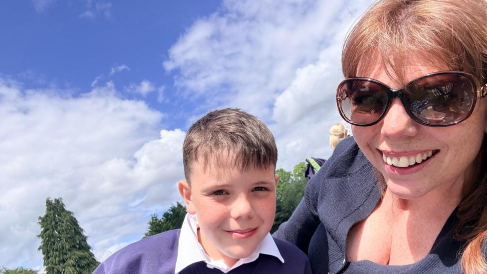 Adam and Karen smiling at the camera. Adam is wearing a white shirt and purple jumper. Karen is wearing a blue cardigan and sunglasses. They are both smiling and there are trees and blue skies behind them.