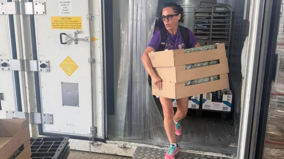 A woman, wearing shorts and a purple T-shirt, with a backpack on her back, walks out of a cold storage unit holding three boxes of food. She has dark hair tied back and is wearing glasses.