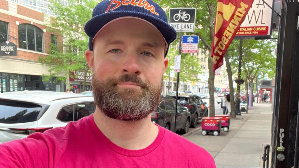 Josh Robertson, wearing a pink top on a Chicago street, with two delivery robots in the background