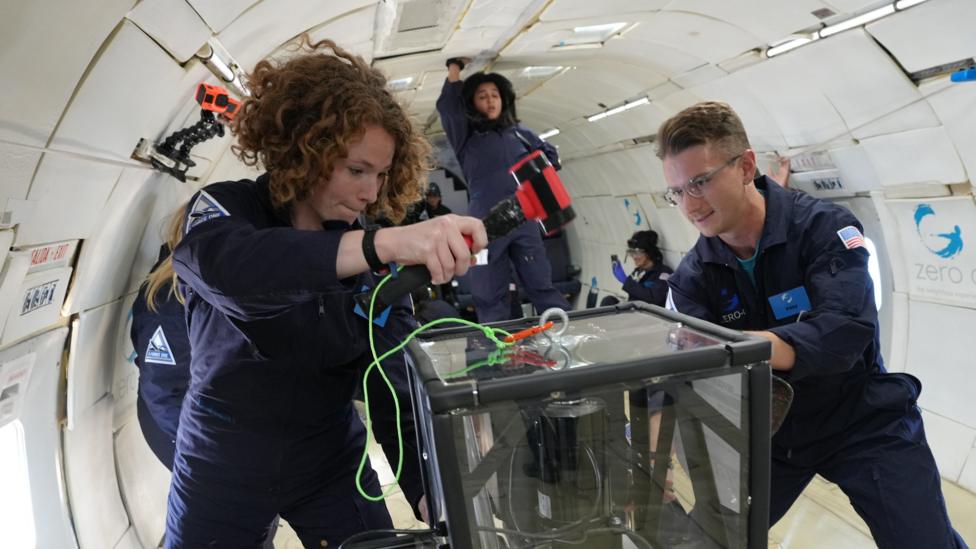 Two young researchers in blue jump suits work on some equipment on a low-gravity flight.