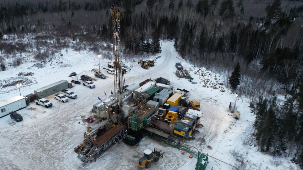 A drilling rig surrounded by snow, woods and industrial buildings and equipment