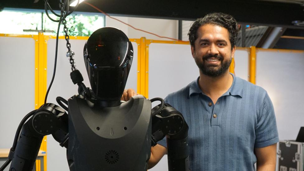 Wearing a blue t-shirt, Sankaet Pathak stands next to the black Phantom robot.