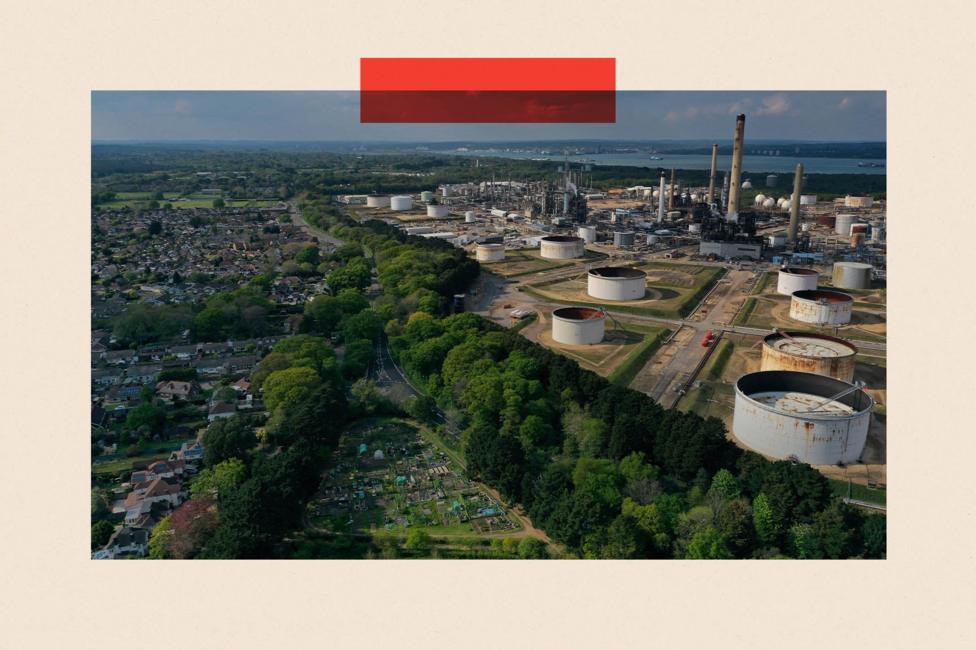 Aerial view of a large oil refinery with chimneys and circular storage tanks, bordered by woodland and a nearby residential neighbourhood, with water visible in the distance; a red rectangular graphic overlays the top of the image.