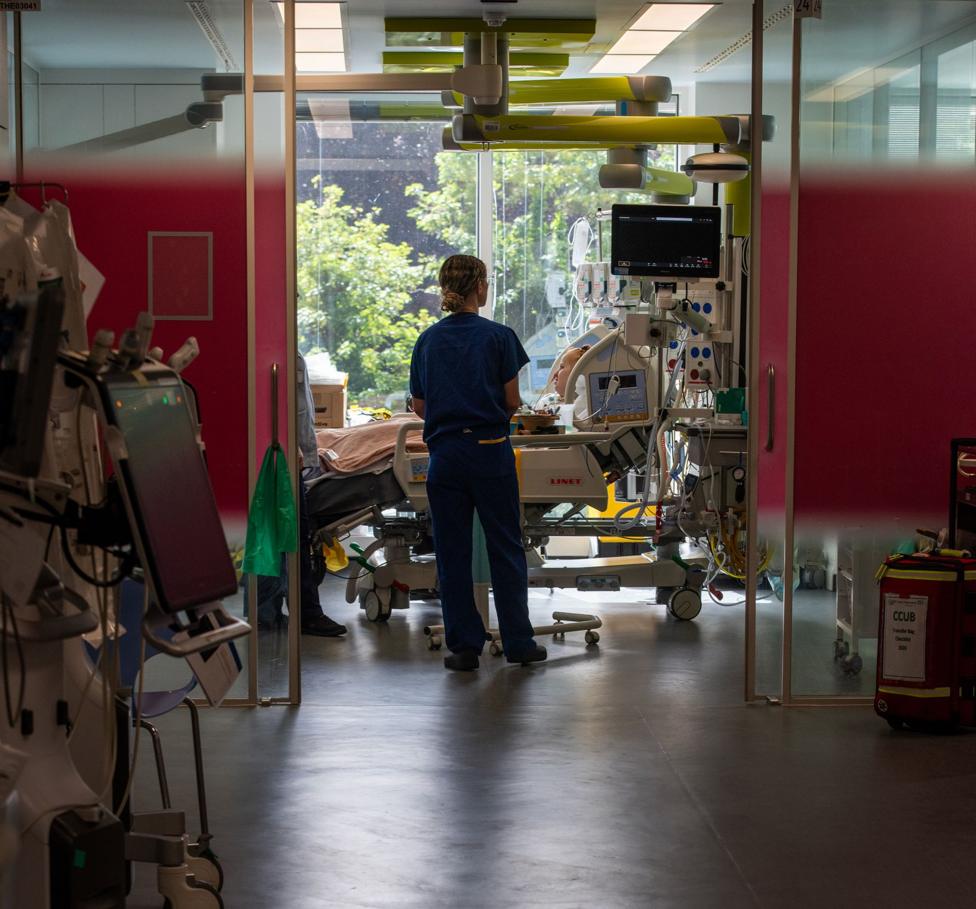 Hollie, 29, is propped up on pillows in her hospital bed in an ICU room inside King's College Hospital, wearing a hospital gown with tubes attached to her nose, and her mother sits by her bedside (unseen) while a nurse checks her.